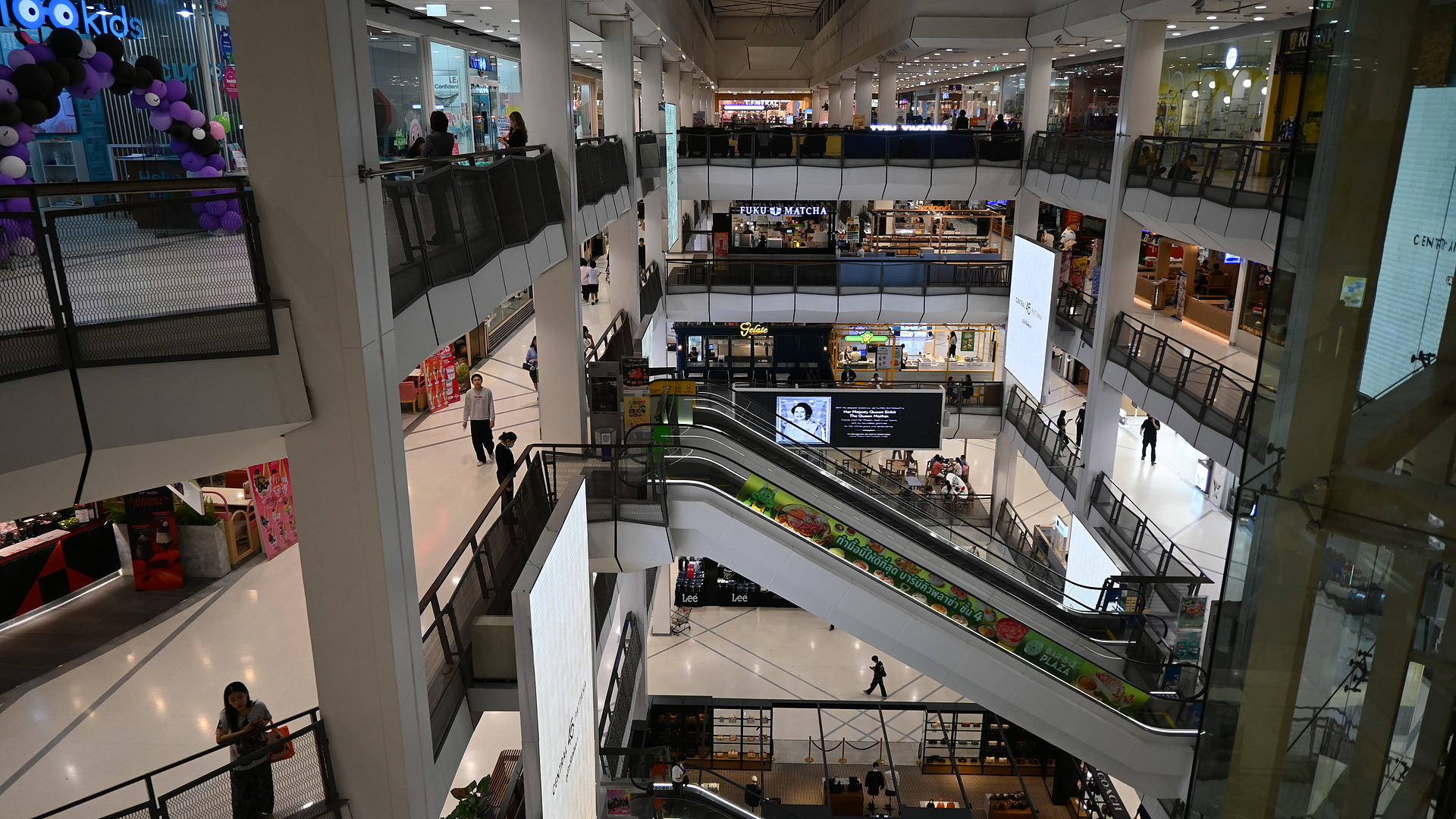 View from the upper escalator inside Grand Arcade Cambridge looking down at shoppers walking, multi-level retail shops, and glass skylight roof